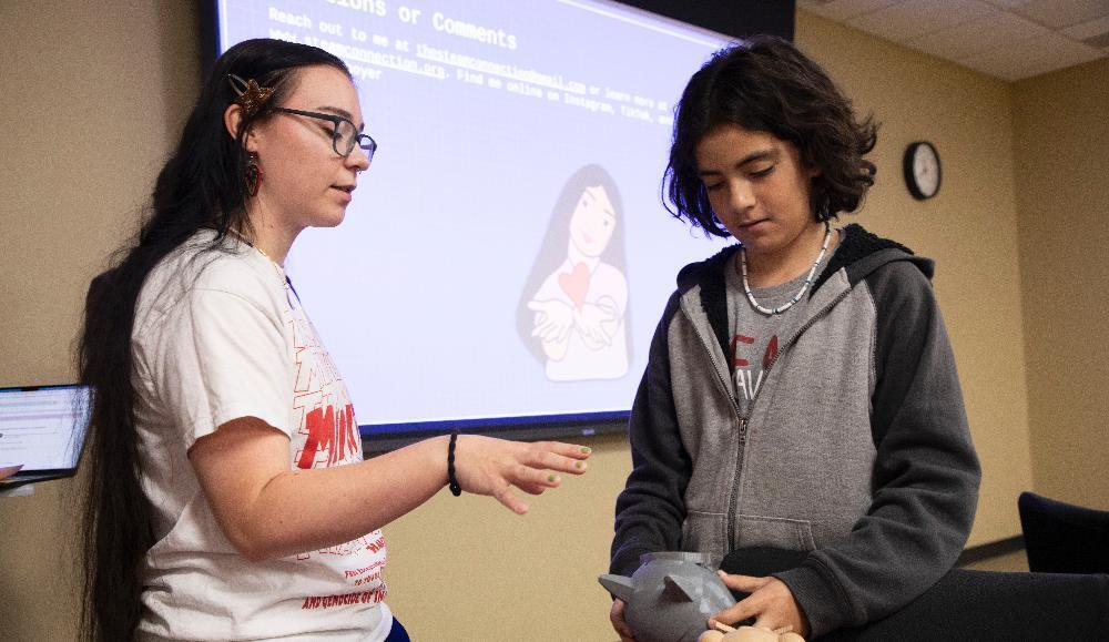 A woman with long dark hair and glasses gestures while speaking to a younger person. The younger person, in a grey hoodie, looks down at a small grey object they are holding. A large screen behind them displays text and an illustration of a person with outstretched hands.