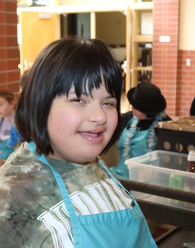 A smiling young person with short dark hair, and a light blue apron over a tie-dye shirt looks at the camera. Other people and a classroom setting are visible in the blurry background.