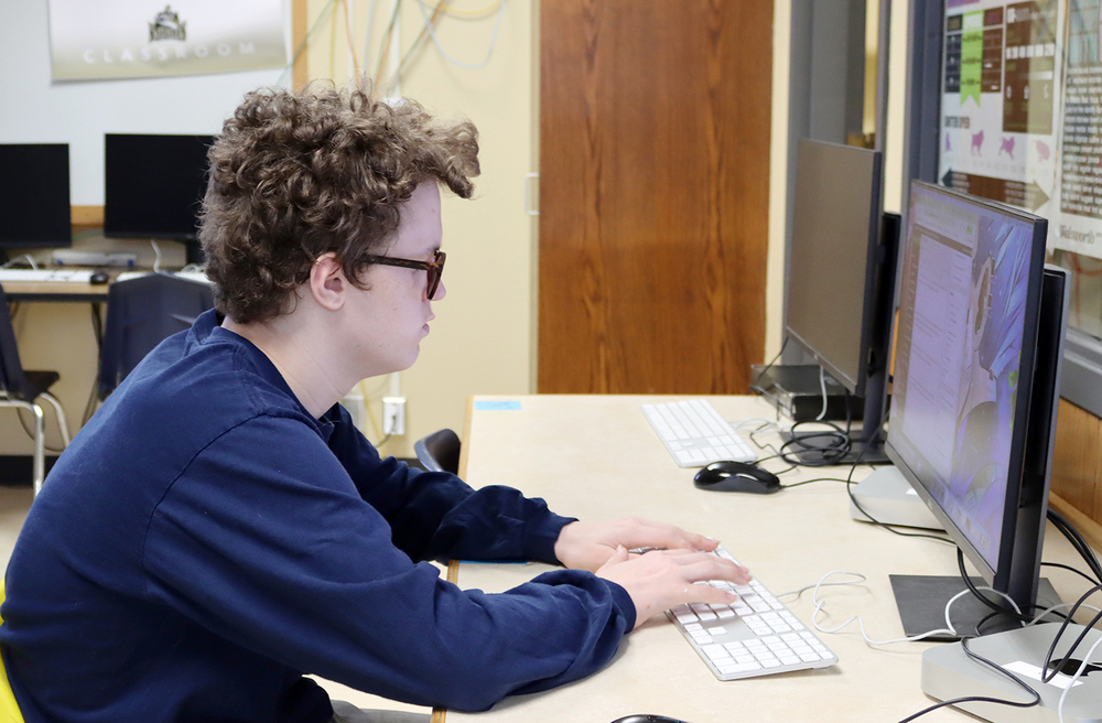 A Southeast High School student works on a newspaper story on his desktop computer. He is typing on a keyboard and is facing left to right.