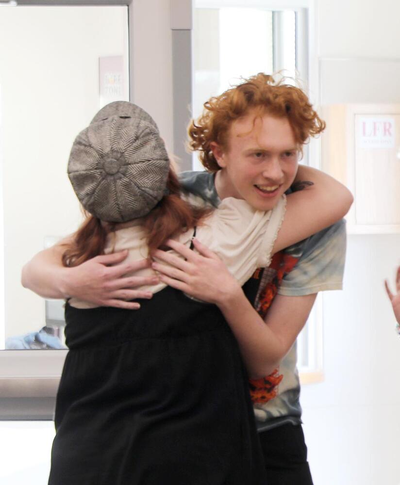 A young person with red hair hugs another person from behind in an indoor hallway. The person being hugged wears a dark dress and a newsboy cap. A portion of a sign reading "LFR" is visible.