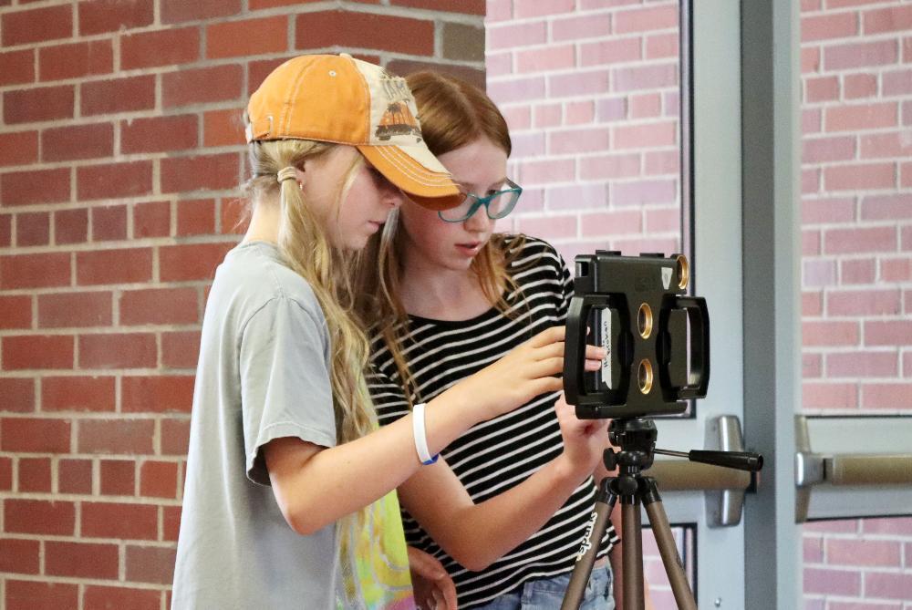 From left, Spark Middle School Tech Camp students Addie and Vera look at a video camera that is mounted horizontally on a tripod in a hallway at Holmes Elementary School. Addie is wearing a t-shirt and a hat that says the word Jamaica on it. Vera is wearing glasses and a short-sleeved shirt.