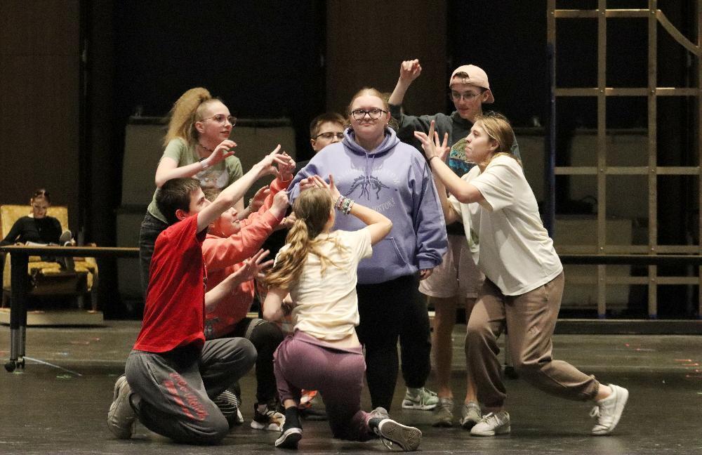 A group of young people, centered around a girl in a purple sweatshirt, are actively performing a scene on a stage. They are interacting physically, with some reaching towards the central figure.