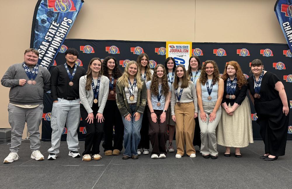 Fourteen smiling students, wearing medals, pose proudly at the 2026 State Journalism Championships. They stand before a black and blue event banner, celebrating their achievements.