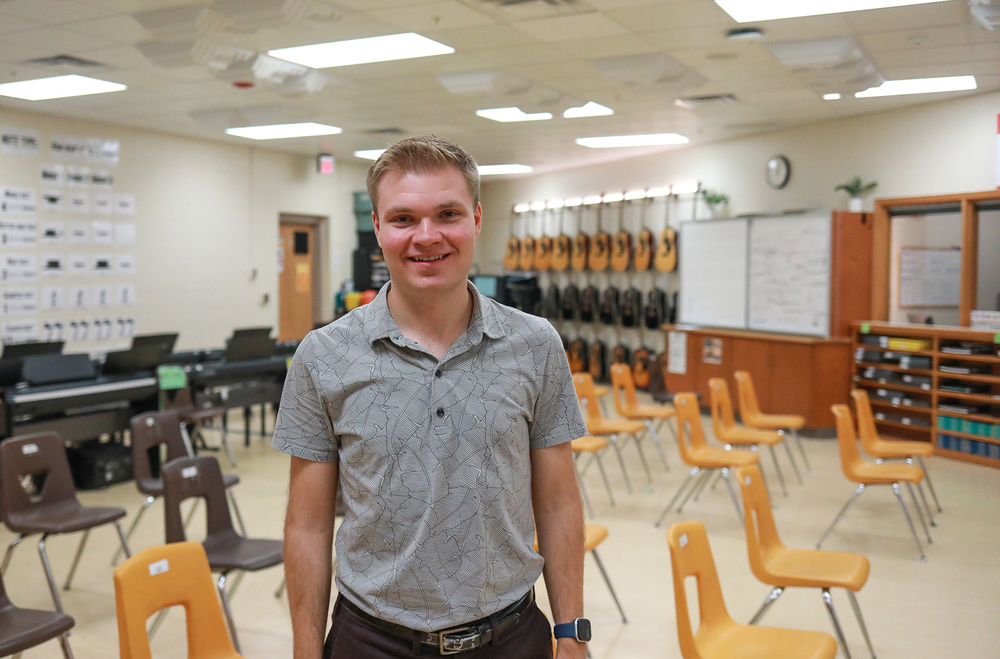 Austin Oltman smiles while standing in his music classroom at Goodrich Middle School. There are more than a dozen chairs on the floor and shelves in the background. There are also three rows of guitars hanging on the wall in the background.