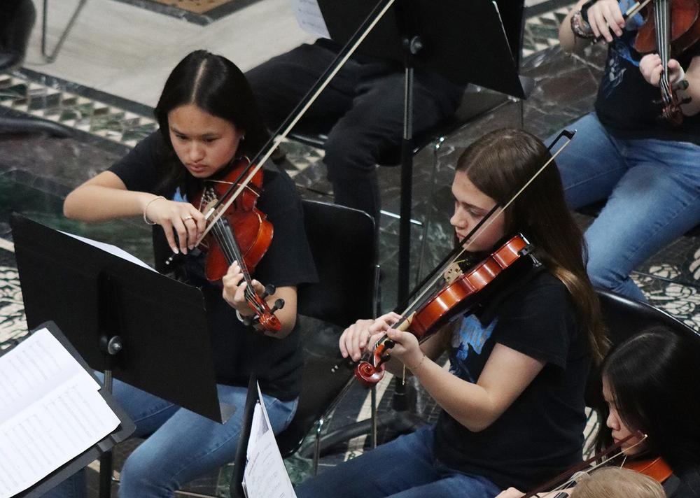Two Schoo Middle School students play their violins in the rotunda of the State Capitol during an orchestra concert. They are holding their instruments with their left hands and their bows with their right hands.