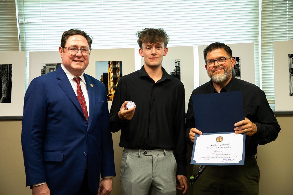 Three smiling men pose. The man on the left wears a blue suit. The young man in the middle holds a white medallion. The man on the right displays a "Certificate of Congressional Recognition" in a blue folder. Framed photos hang behind them.