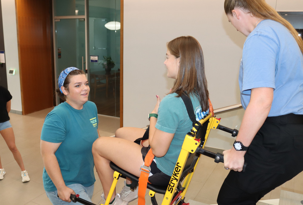 Two students practice carrying a classmate up a stairway in their emergency medical technician class. One girl is sitting in the chair lift and is facing right to left. One girl is holding handles at the bottom of the chair lift, and the other girl is holding handles at the back of the chair lift.