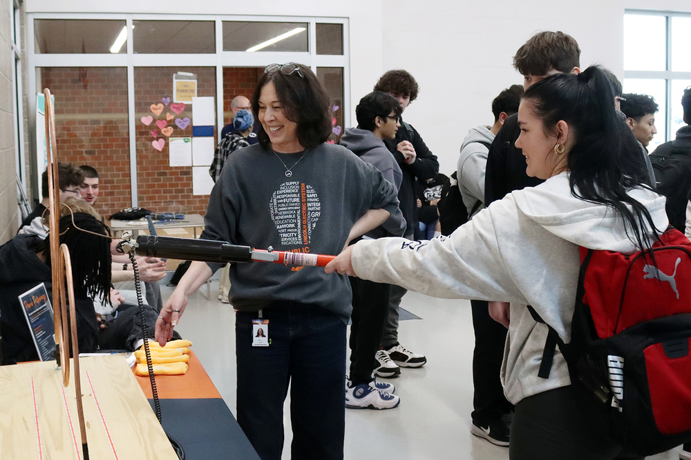 From right to left, a Lincoln North Star student holds an electric rod that has a circular coil at the end. She is moving the circle over a loop without having the circle touch the loop. An electrical representative is smiling in the middle of the photo with other students in the background.