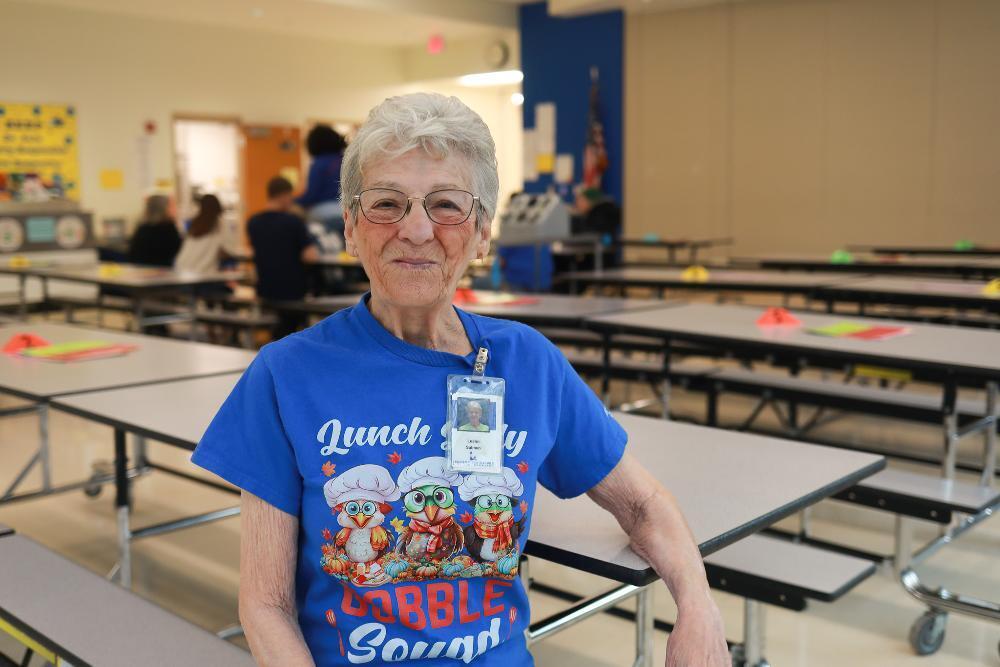 A woman with short gray hair and glasses smiles in a school cafeteria. She wears a blue "Lunch Lady Gobble Squad" T-shirt and a name badge, standing beside long cafeteria tables with a few people blurred in the background.