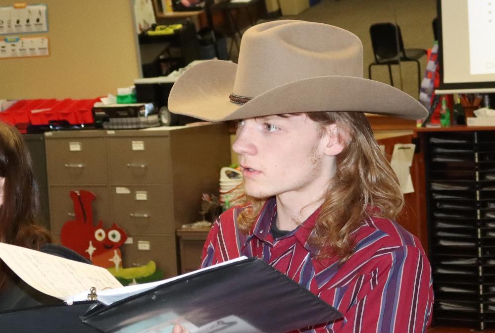 Northeast High School senior Tayler performs with the Chamber Singers group in the choir room. He is wearing a cowboy hat and is holding a songbook while he is singing.