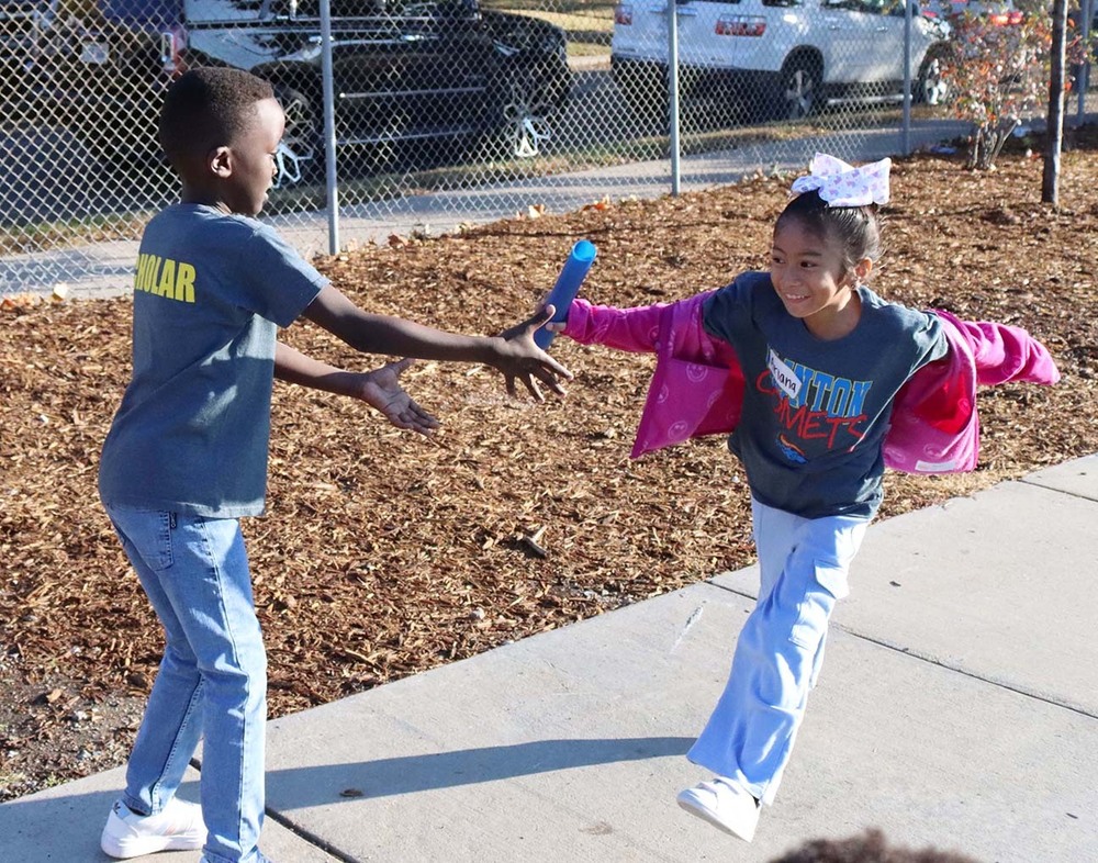 A Clinton Elementary School student hands a baton to a classmate during a relay race on a sidewalk by the school's playground.