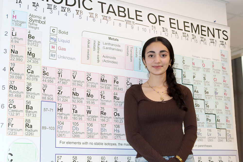 International Baccalaureate Focus Program senior Khalidah smiles in front of a large poster of the periodic table of elements. The poster is taped to the wall of a chemistry classroom.