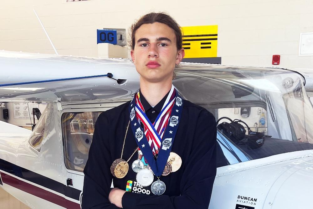 A young man, wearing several medals, stands arms crossed in front of a small airplane. The airplane is light-colored with dark accents. "DUNCAN AVIATION" is visible on its side.