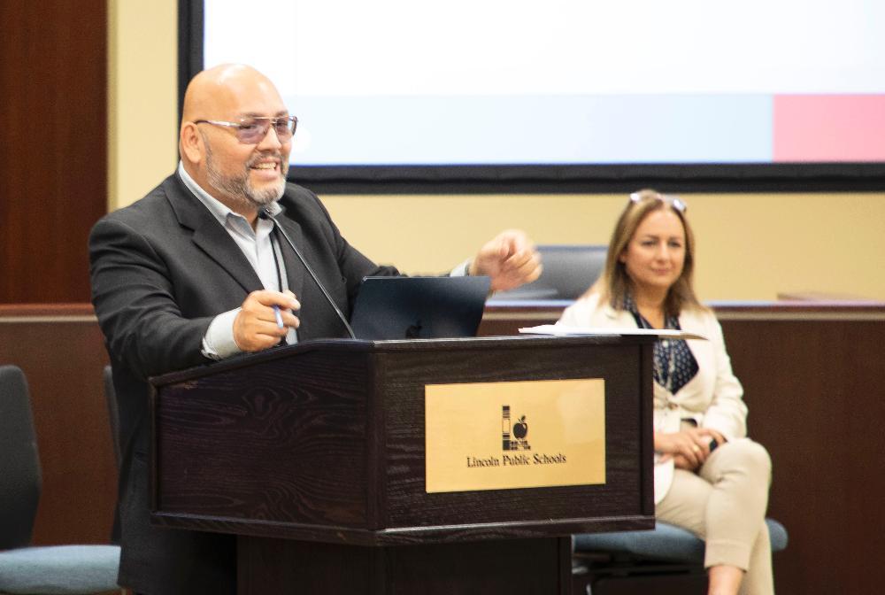 LPS Federal Programs representatives Oscar Rios Pohirieth and Raya Al Zuhair engage with the audience at a program in the Steve Joel District Leadership Center. Rios Pohirieth is smiling and motioning at a podium in the center of the picture, and Al Zuhair is sitting in a chair behind him on the righthand side. There is a small section of an informational screen on the upper-righthand side of the picture.