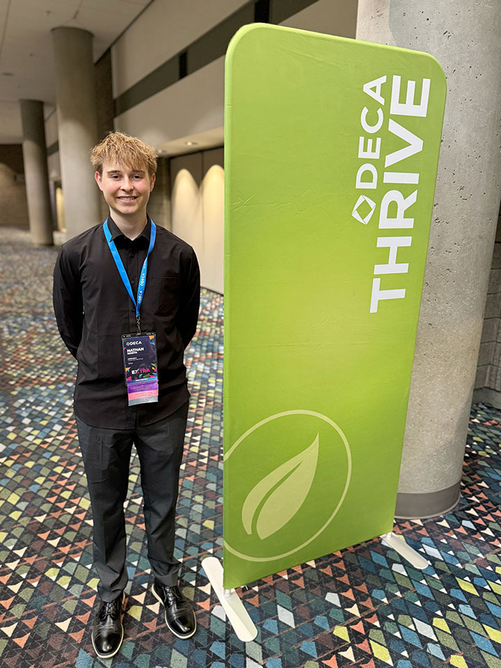 East student Nathan North stands next to a large rectangle-shaped sign at the DECA International Career Development Conference. The sign has a symbol of a leaf and the words DECA Thrive on it.