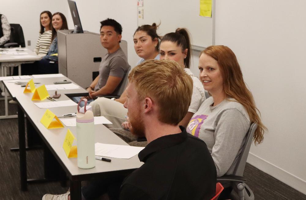 Several people are seated in a classroom. A red-haired man sits with his back to us, beside a smiling woman with long hair. Tables in front of them hold papers, pens, and yellow name cards, suggesting a training session.