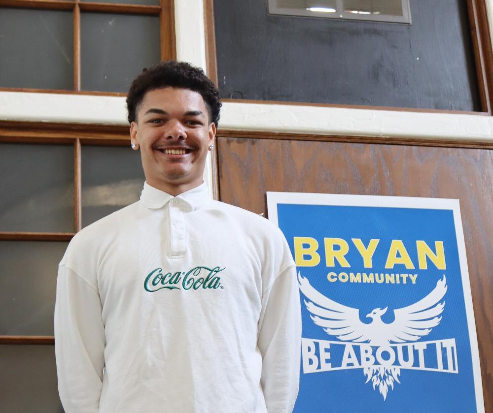 A young man, smiling, stands before a "Bryan Community" poster featuring a phoenix. The poster reads, "BRYAN COMMUNITY BE ABOUT IT". He wears a white Coca-Cola shirt.