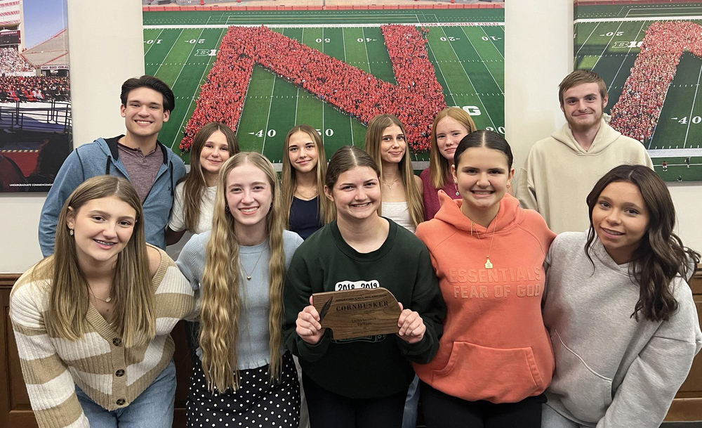 Eleven Southwest High School students smile in two rows. There are five girls in the first row and four girls and two boys in the second row. The girl in the middle of the first row is holding a Cornhusker Award plaque.