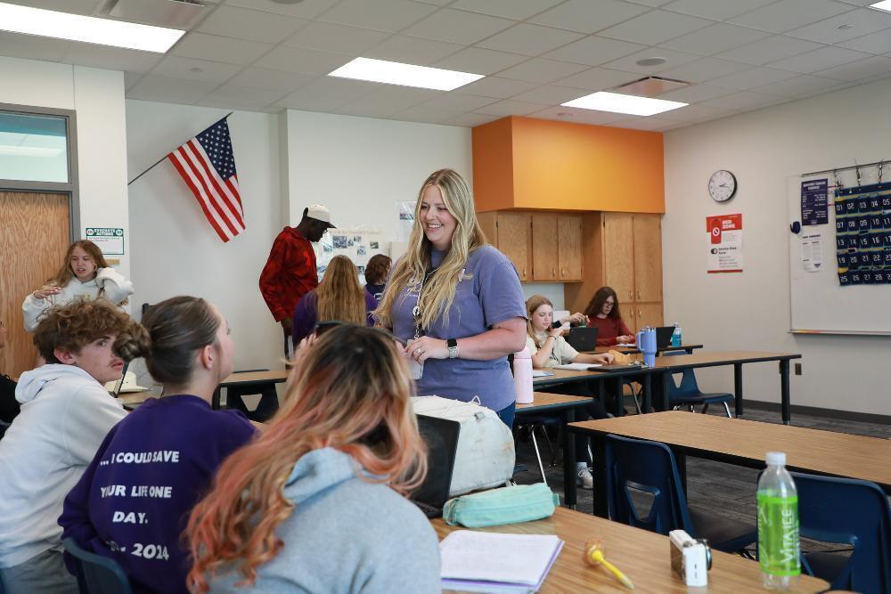 Lindsay Pierce smiles while she teaches a social studies class at Northwest High School. She is standing in between several two-person desks that are in the classroom. She is speaking to a girl who is sitting in front of a laptop computer.
