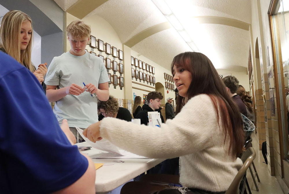 A Northeast High School student sits at a table and passes a piece of paper to two students standing in front of the table. They are in the main hallway of the high school.