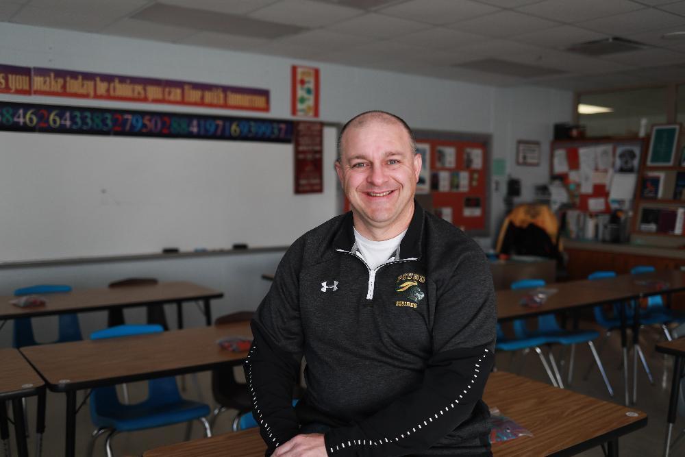 A man sitting inside a classroom wearing a black shirt.