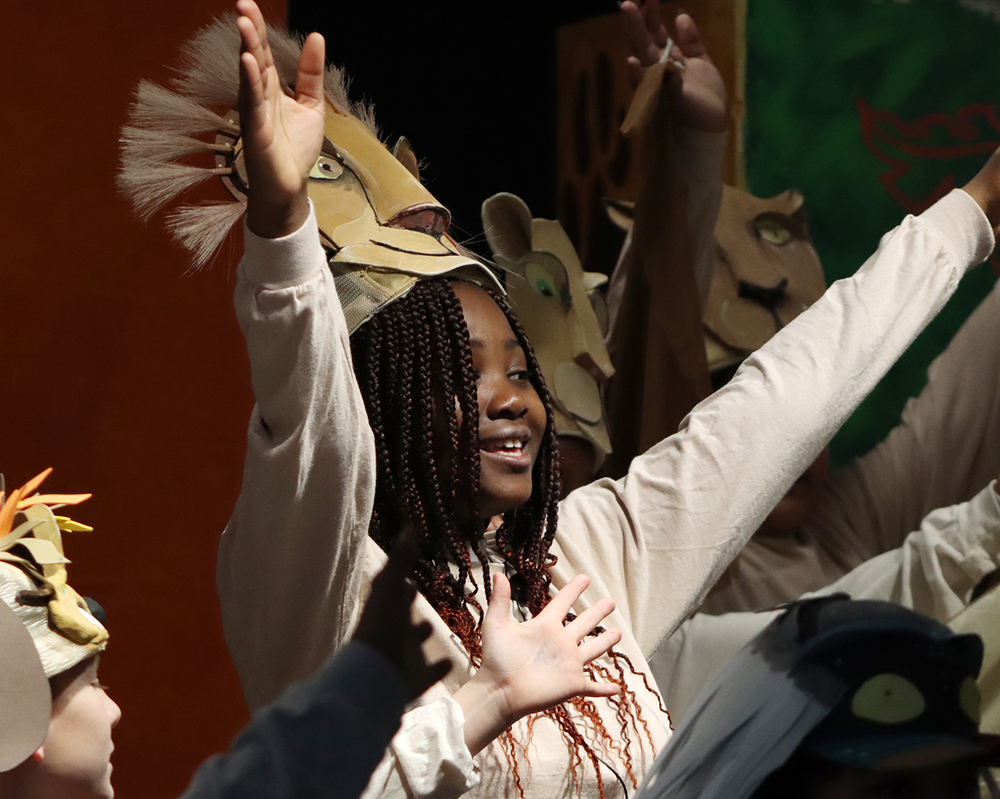 A Belmont Elementary School student raises both of her hands while singing "Circle of Life" in The Lion King. She is portraying the main role of Simba and is wearing a lion costume.