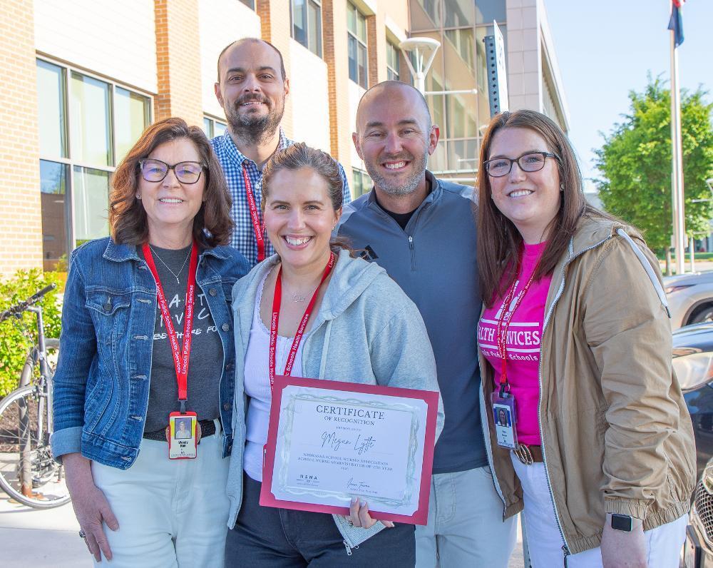 Five smiling adults, three women and two men, pose outside a building. The woman in the middle holds a "Certificate of Recognition" for Megan Lytte. Several wear Lincoln Public Schools Health Services lanyards, suggesting a team celebration.