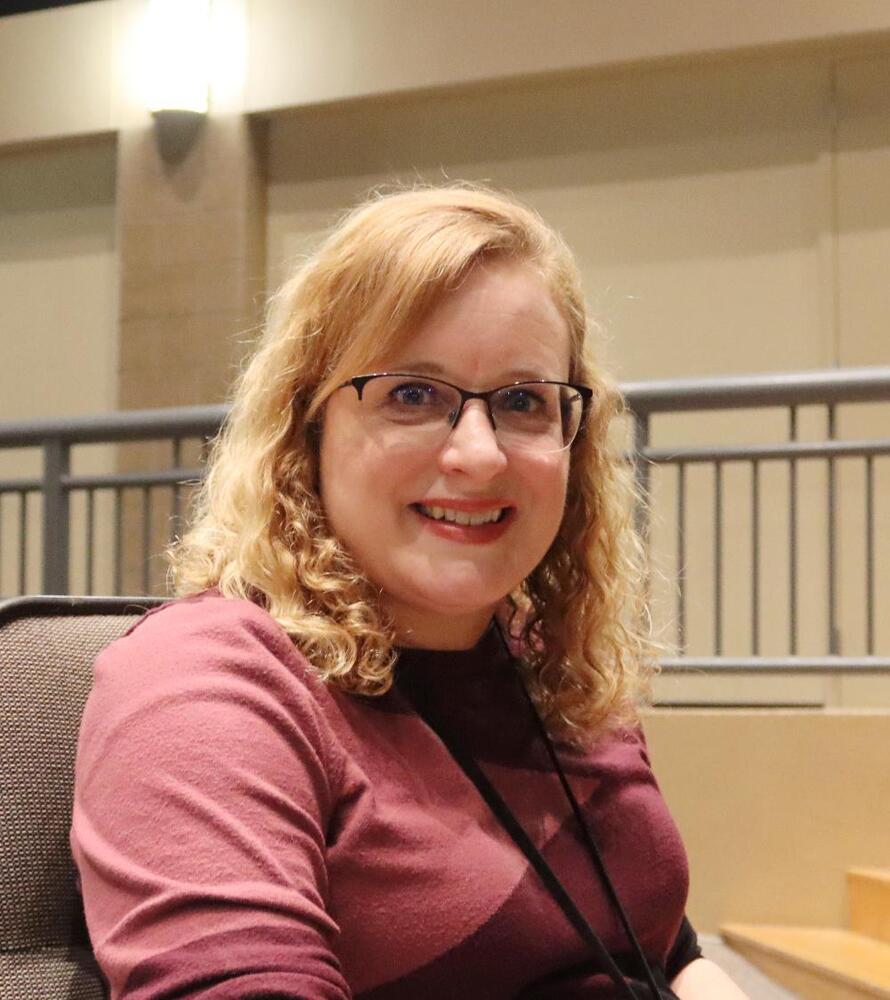 A woman with curly blond hair and glasses sits in a burgundy sweater. She is smiling, in a building with a light-colored wall and a metal railing.