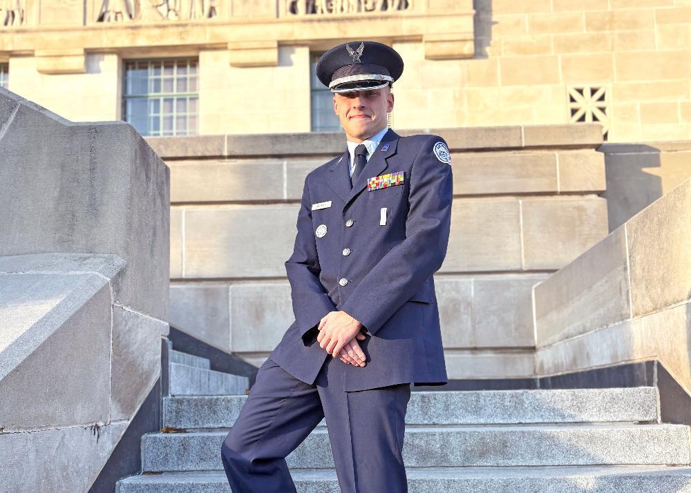 A man in a dark blue Air Force dress uniform stands with hands clasped on a stone staircase. The background features a large, light-colored stone building.