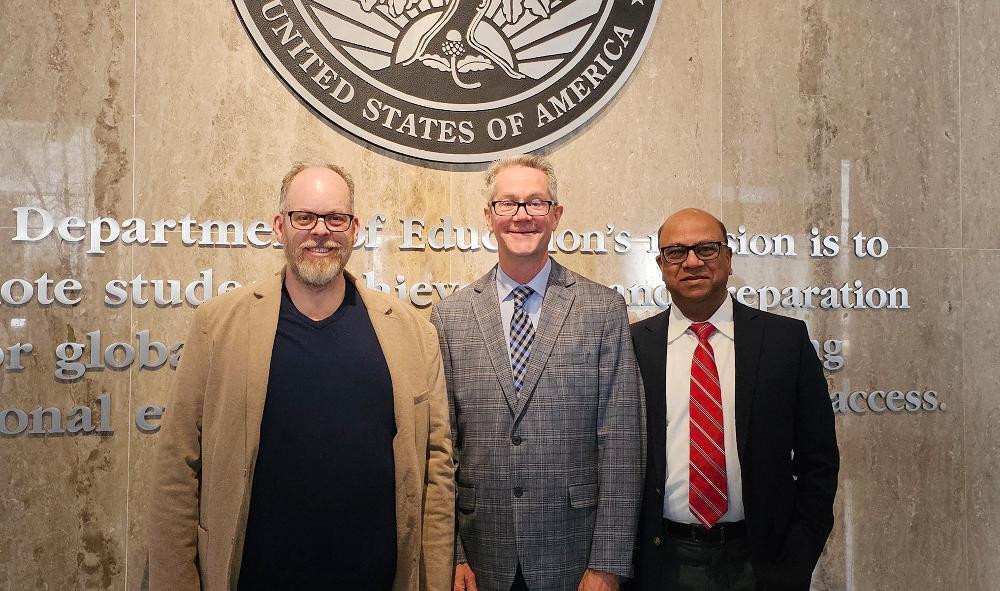 Three men stand posing, in front of a Department of Education wall.