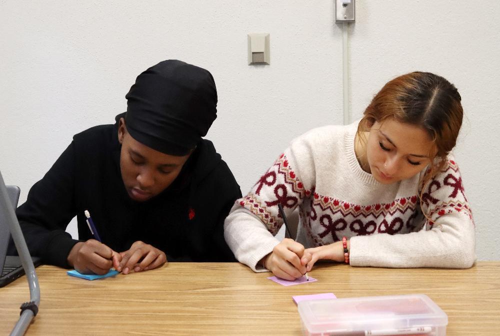 From left, Bryan Community students Shy and Bel write information about scholarship letters during a class activity. Both are using their right hands to put information on Post-It notes while they are sitting at a table. Shy is wearing a sweatshirt and Bel is wearing a knitted sweater.
