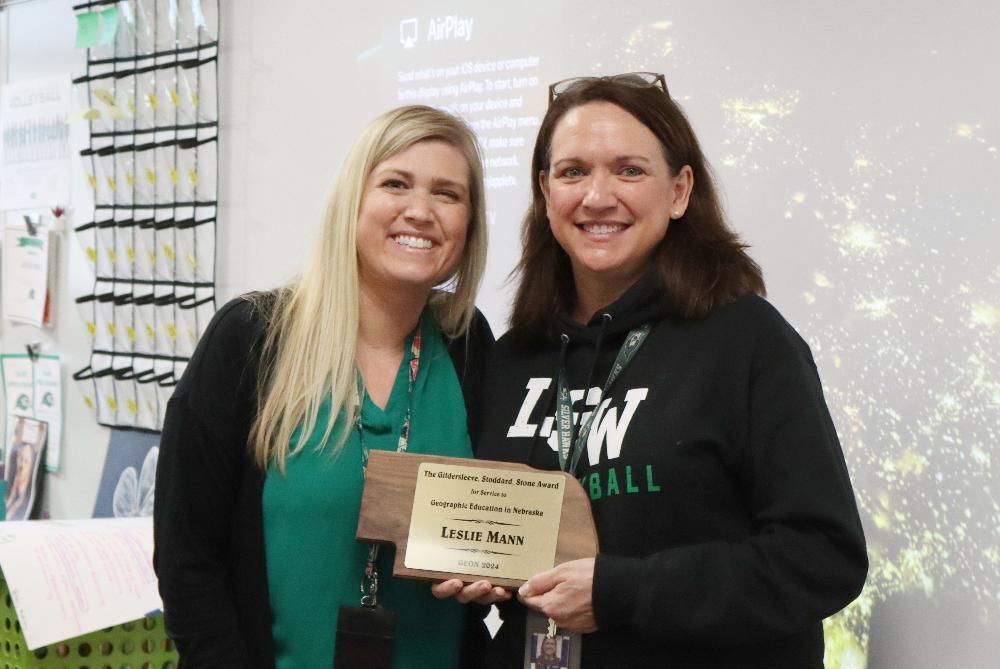 Two women hold a wooden award plaque. The plaque reads: "The Gildersleeve, Stoddard, Stone Award for Service to Geographic Education in Nebraska LESLIE MANN GEON 2024". They stand in a classroom.