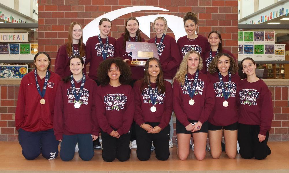 Thirteen smiling young women, wearing maroon "GATORS" shirts, proudly display medals and a "State Champions" basketball trophy. They pose in two rows, some kneeling, celebrating their victory against a brick wall background.