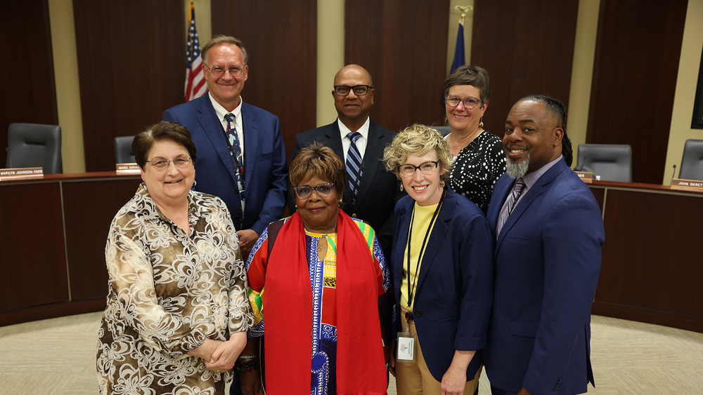 Seven smiling adults, four women and three men, stand together in the Lincoln Public Schools board room. They are professionally dressed, with United States and Nebraska state flags visible in the background.