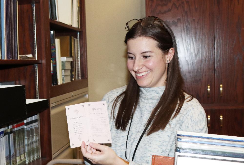 Sara Scott smiles as she displays a program from Lincoln High's 1883 commencement ceremony. She is holding the small program just under her right shoulder and is looking down at it. She is standing in a special archives room that features rows of books and programs on the lefthand side of the picture. There are wooden cabinets filled with additional items behind her.