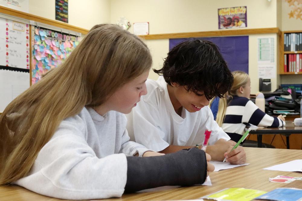 From left, a girl and a boy are sitting at a table in a math classroom at Irving Middle School. The girl has a soft cast on her right arm and is looking left to right. She is holding a pen in her right hand. The boy is smiling and is writing on a piece of paper with his left hand. They are taking part in an escape room activity to prepare for a math test about subjects such as polynomials, fractions and decimal-based division.