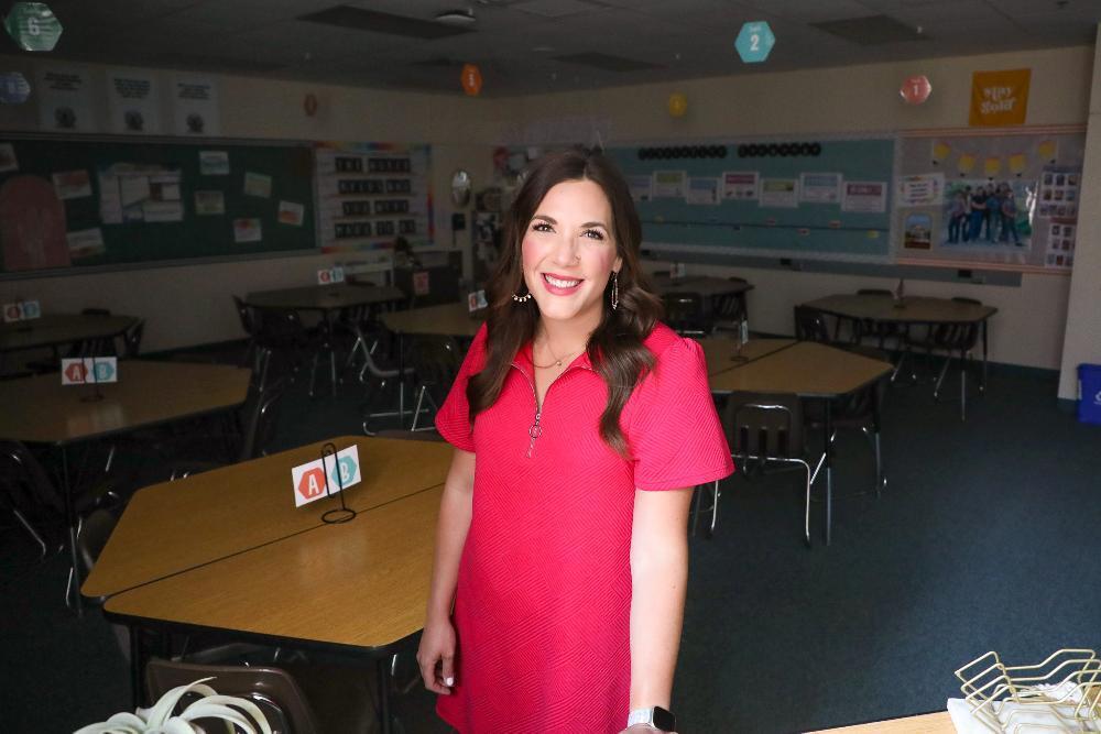 A woman in a pink dress stands in a classroom. Multiple tables and chairs are arranged. The room has colorful decorations and learning materials on the walls.