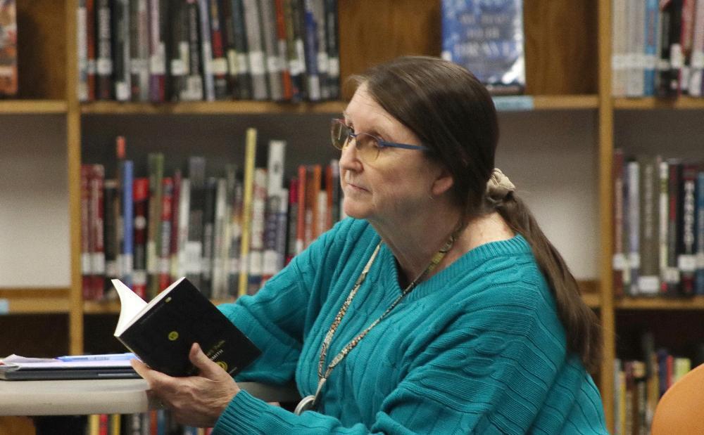 A Mickle Parent-Teacher-Student Organization member sits at a table in the middle school's library. She is wearing a sweater and is holding a book open with both hands. There are full bookshelves in the background.