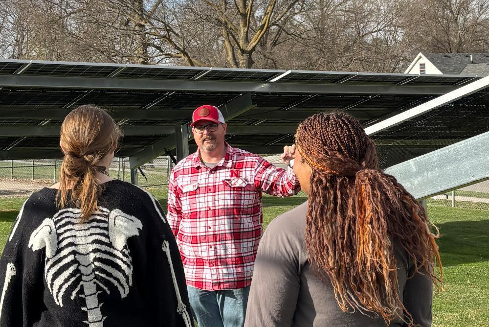 A man in a red plaid shirt talks to two women outdoors. One woman wears a skeleton sweater. Behind them, rows of solar panels span a grassy area under a bright sky, with trees and a house in the distance.