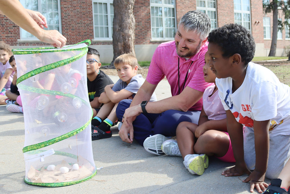 Randolph Elementary School Principal Ryan Erickson smiles as he sits in a row of eight first graders on a sidewalk outside the school building. They are preparing to watch a butterfly get released from a cylinder butterfly container.