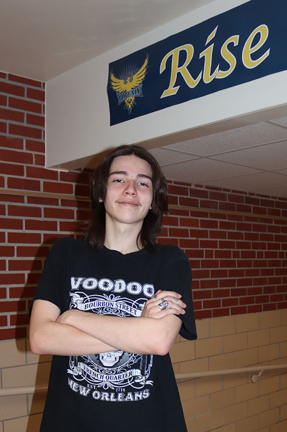 Bryan Community senior Tommy is standing with his arms folded in a hallway. He is standing under a beam on the ceiling that has a sign with the word 'Rise' on it.