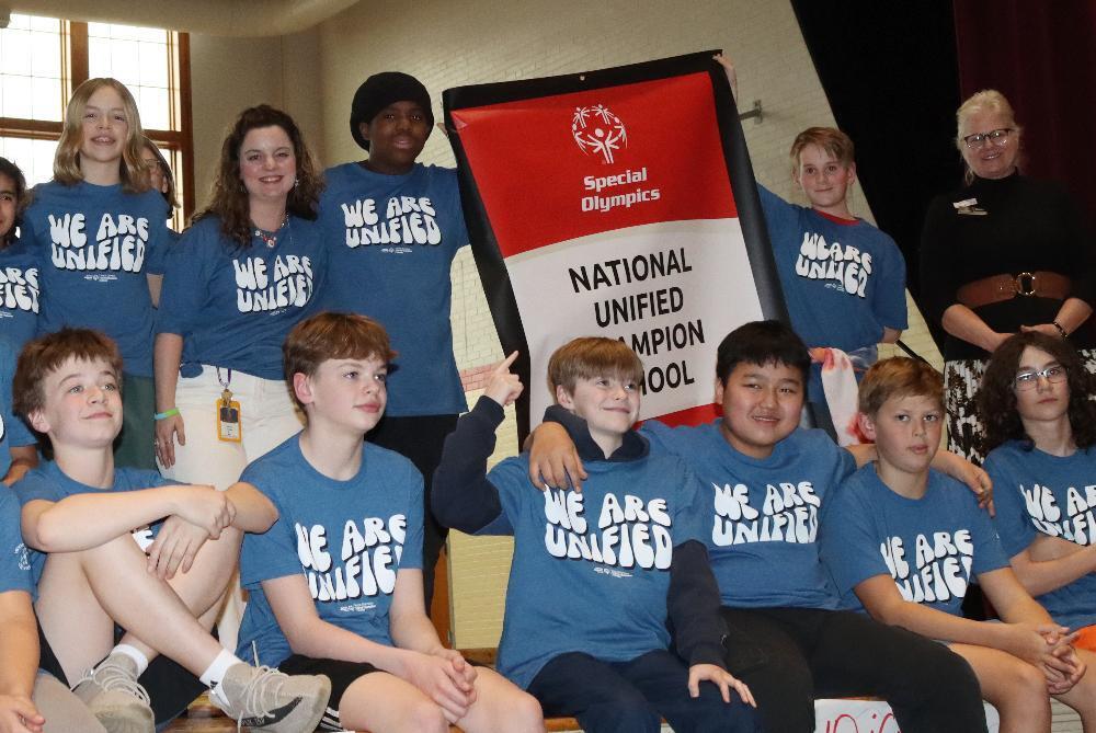 A diverse group of 14 people, mostly students, wear blue "WE ARE UNIFIED" shirts. They pose smilingly, holding a red and white Special Olympics banner declaring them a "NATIONAL UNIFIED CHAMPION SCHOOL."