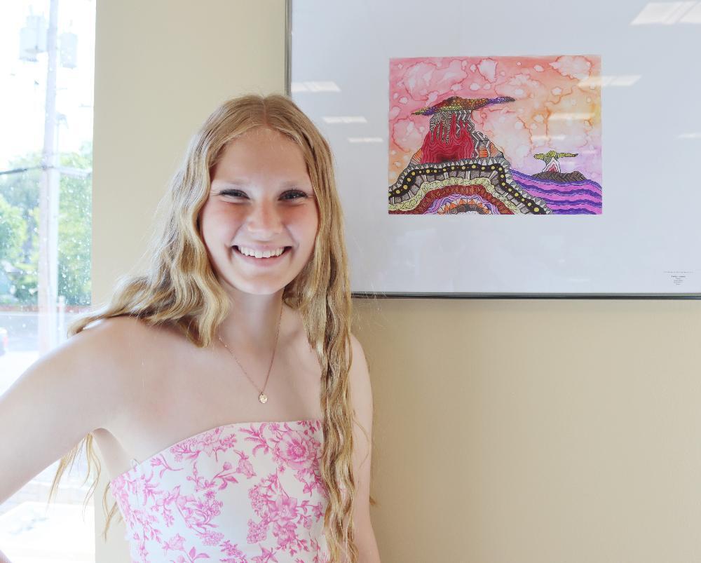 A young woman with long blond hair, wearing a pink floral strapless dress, smiles beside an abstract watercolor painting of stylized mountains and sky.
