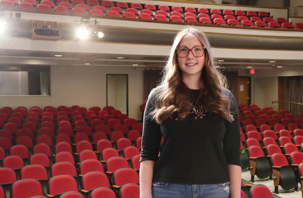Lincoln High senior Soren smiles on stage of the school auditorium. Empty rows of seats are behind her and two spotlights are turned on to shine on the stage.