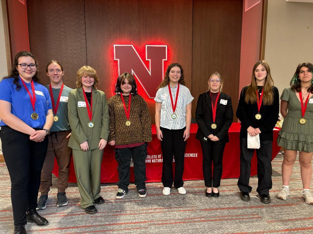 Eight students stand, wearing medals around their necks, in front of a red backdrop featuring a large "N" logo.