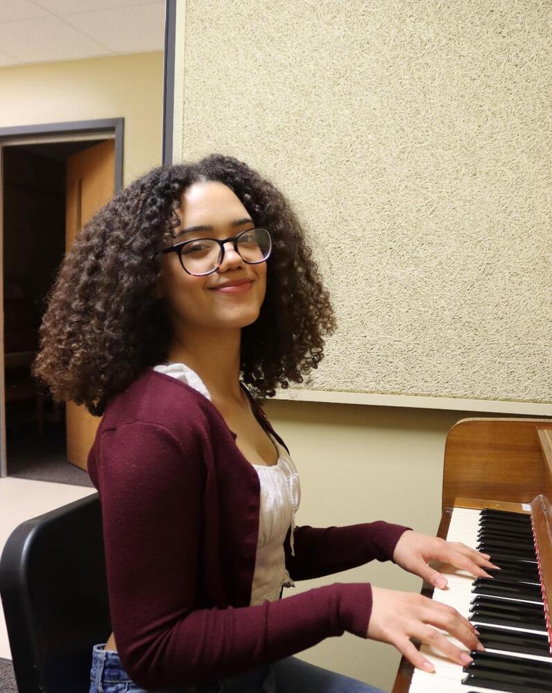 A young woman with curly hair plays a piano in a music room. She wears glasses and a maroon cardigan.