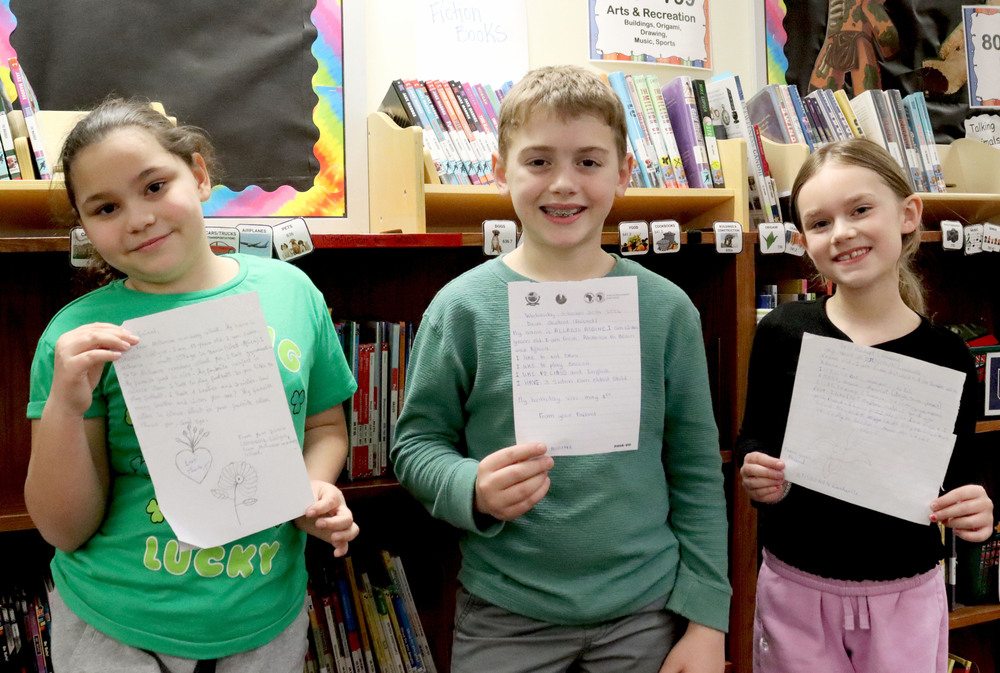 Three Kahoa Elementary School students hold letters they received from pen pals in Benin. They are standing in front of several bookshelves in the school library.