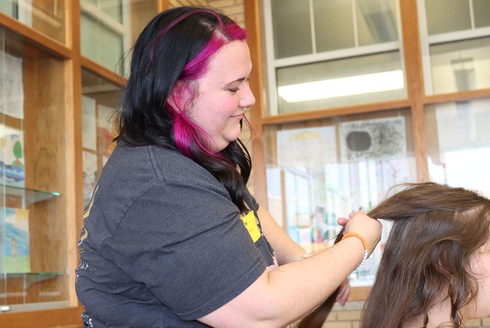 College of Hair Design student Karlee Rademacher smiles as she works on styling the hair of a Lincoln High student at the inaugural Homecoming preparation event. She is facing left to right and is holding a large strand of the student's hair with both hands.