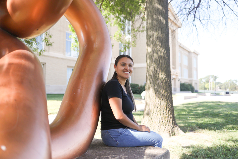 Woman sitting beside a large outdoor sculpture on a university campus, smiling toward the camera. Trees and a campus building are visible in the background.