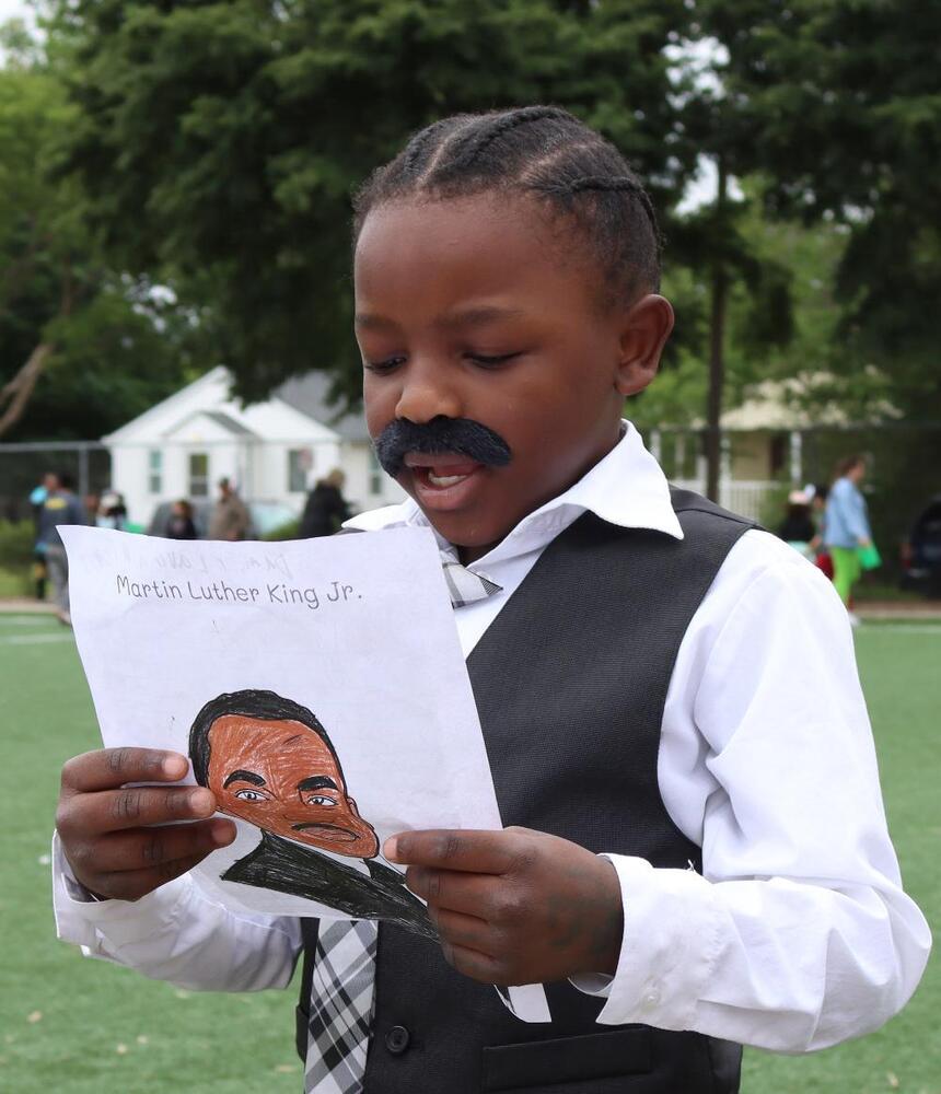 A boy displays a drawing of Martin Luther King Jr., outdoors on a grassy field. "Martin Luther King Jr." is written at the top of the drawing.